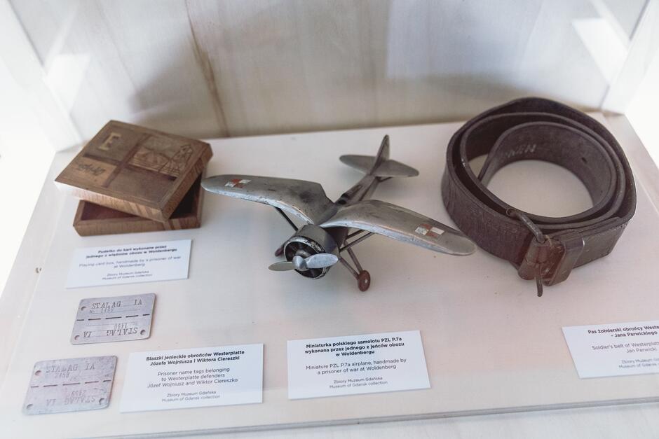 Na zdjęciu znajdują się przedmioty z Muzeum Gdańska, w tym drewniane pudełko na karty do gry, miniaturowy model samolotu PZL P.7a oraz skórzany pas wojskowy. Każdy przedmiot ma opis informujący o jego pochodzeniu, w tym m.in. o tym, że zostały wykonane przez jeńców wojennych.