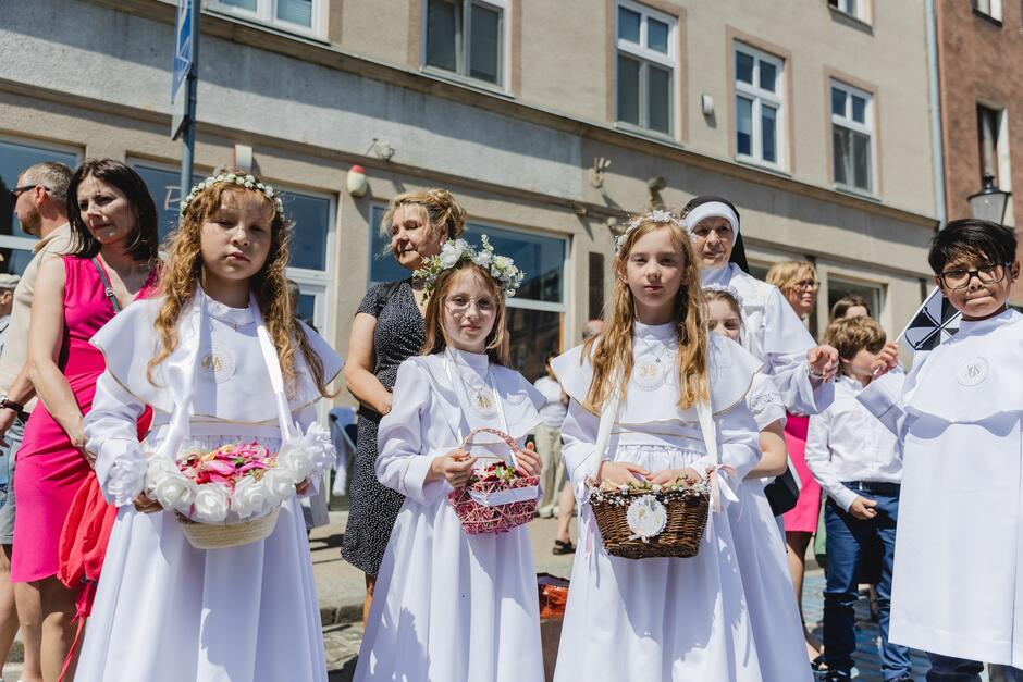 Dziewczynki w białych sukienkach, z koszyczkami, w których znajdują się płatki kwiatów