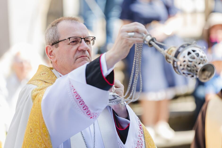 Mężczyzna w szatach biskupich, podczas uroczystości. Rękami kołysze kadzielnicę, która zawieszona jest na łańcuchu  