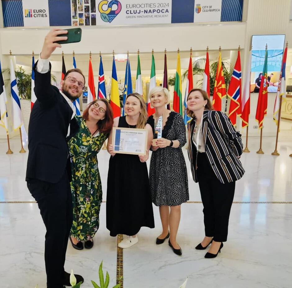 Na zdjęciu grupa pięciu osób robi sobie selfie podczas ceremonii wręczenia nagród. Trzymają w rękach dyplom oraz statuetkę, stojąc na tle szeregu flag różnych krajów, co sugeruje międzynarodowy charakter wydarzenia.