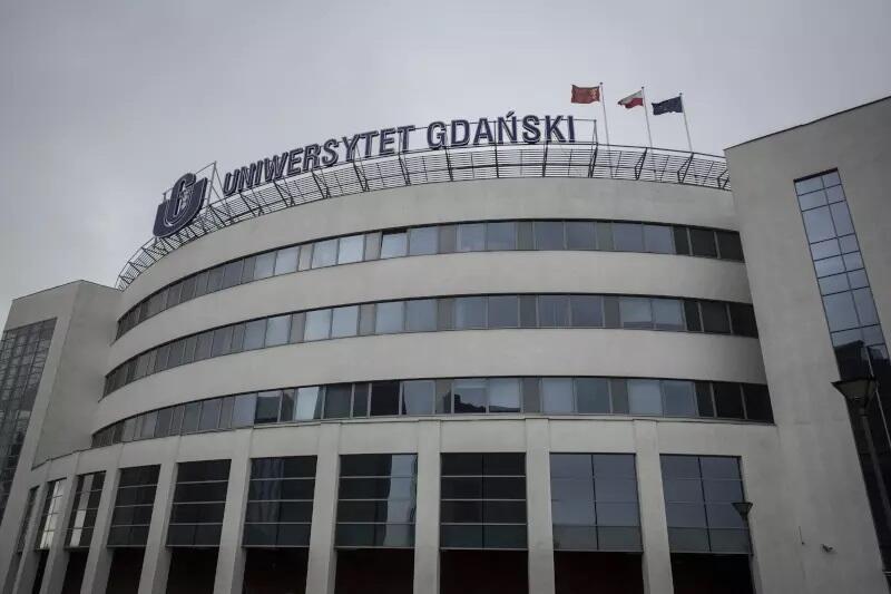 Na zdjęciu widzimy nowoczesny budynek Uniwersytetu Gdańskiego. Na szczycie budynku znajduje się duży napis "Uniwersytet Gdański" oraz trzy flagi powiewające na masztach, w tym flaga Polski