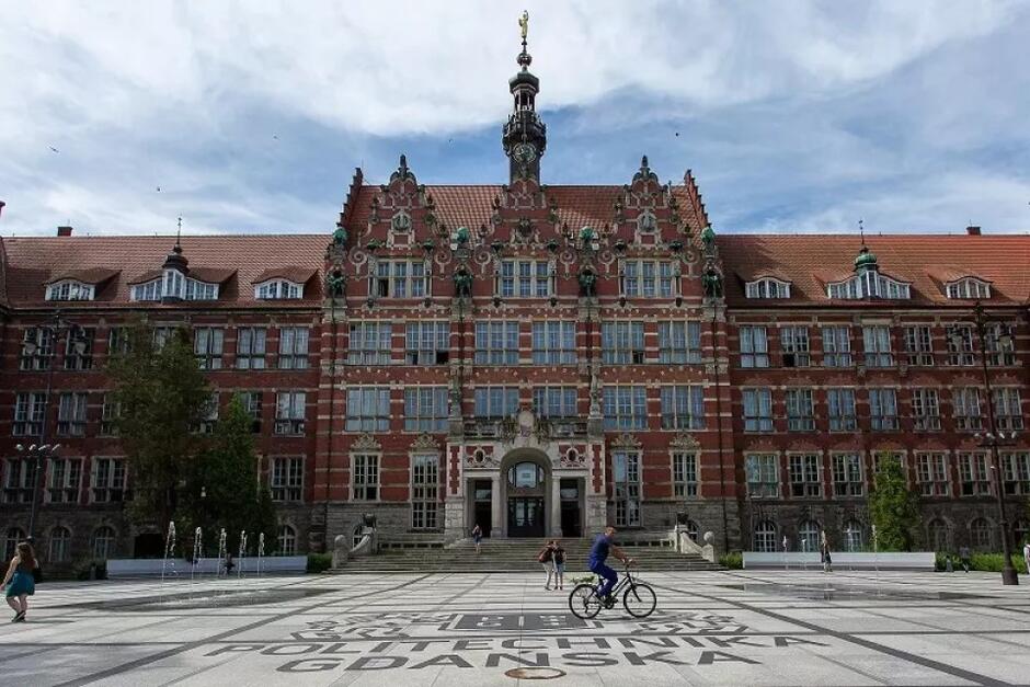 Na zdjęciu widzimy główny budynek Politechniki Gdańskiej, charakteryzujący się zabytkową architekturą z czerwonej cegły i licznymi zdobieniami. Przed budynkiem znajduje się duży plac, na którym widoczne są napisy "Politechnika Gdańska", a po nim przejeżdża rowerzysta, co dodaje dynamiki scenie