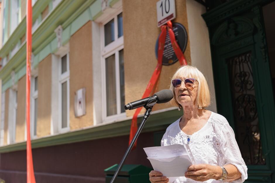 Na zdjęciu znajduje się starsza kobieta w okularach przeciwsłonecznych, przemawiająca do mikrofonu na tle budynku oznaczonego numerem 10. Budynek jest ozdobiony czerwonymi wstążkami, a na jego fasadzie widoczna jest pamiątkowa tablica.