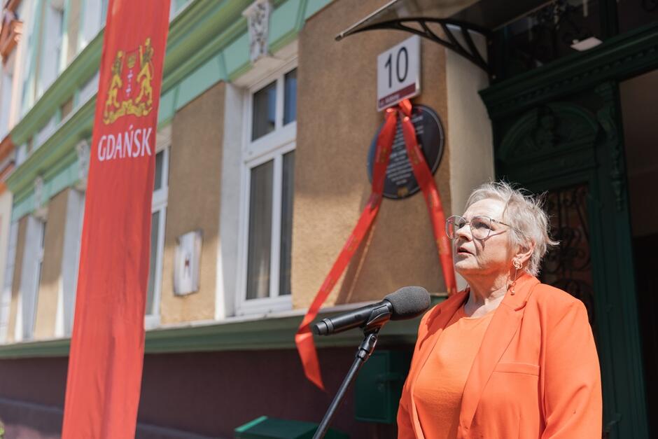 Na zdjęciu widoczna jest starsza kobieta w okularach i pomarańczowym stroju, przemawiająca do mikrofonu na tle budynku oznaczonego numerem 10. Obok niej znajduje się czerwona flaga z herbem i napisem Gdańsk , a na ścianie budynku widać pamiątkową tablicę ozdobioną czerwoną wstążką.