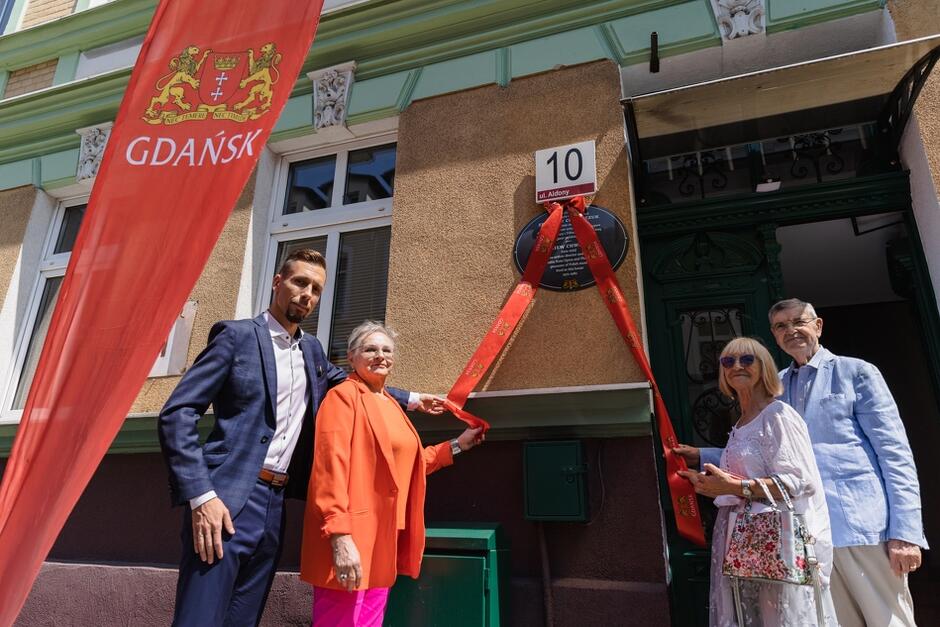 Na zdjęciu widać cztery osoby stojące przed budynkiem oznaczonym numerem 10, ulica Aldony, w Gdańsku, trzymające wstążkę przeciętą podczas odsłonięcia pamiątkowej tablicy. Tablica poświęcona jest Zbigniewowi Chwedczukowi, a w tle widoczna jest czerwona flaga z herbem Gdańska.