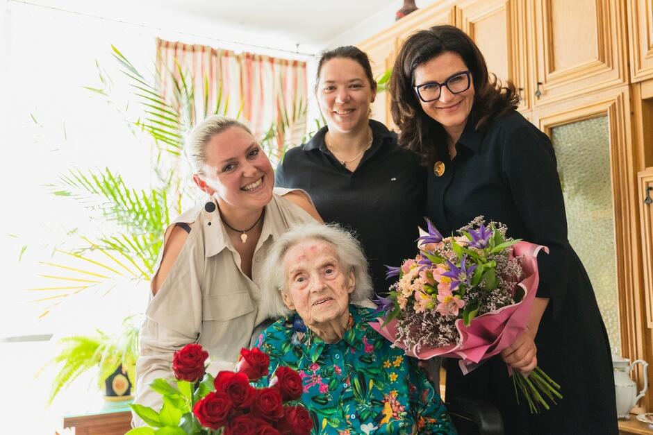 Na zdjęciu widzimy starszą kobietę siedzącą na krześle, otoczoną trzema uśmiechniętymi osobami. Kobieta trzyma bukiet czerwonych róż, a jedna z towarzyszących jej kobiet trzyma bukiet kolorowych kwiatów.