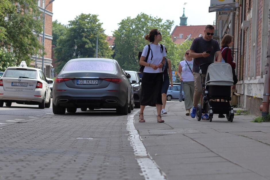 na zdjęciu zbliżenie na fragment asfaltowej jezdni i chodnika, po którym idzie dwoje dorosłych ludzi z wózkiem dziecięcym, to po prawej, a po lewej widać dwa samochody osobowe które się mijają na ulicy