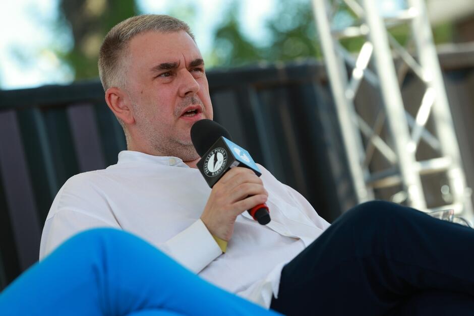 Na zdjęciu znajduje się mężczyzna siedzący na scenie, trzymający mikrofon z logotypem. Ubrany jest w białą koszulę i niebieskie spodnie, a za nim widać elementy konstrukcji scenicznej oraz rozmyte tło