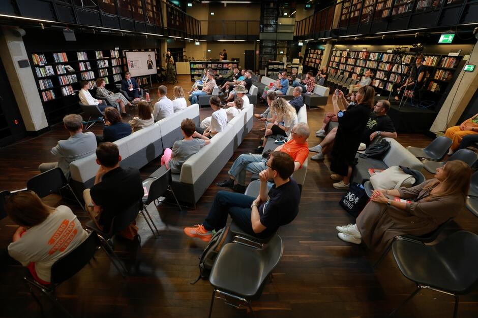 Na zdjęciu widzimy spotkanie literackie w nowoczesnej bibliotece. Uczestnicy siedzą na kanapach i krzesłach, słuchając panelu dyskusyjnego, a półki z książkami w tle tworzą przytulną atmosferę.
