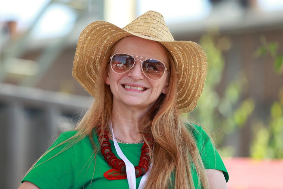 Na zdjęciu znajduje się uśmiechnięta kobieta z długimi rudymi włosami, nosząca duży, słomkowy kapelusz i okulary przeciwsłoneczne. Ma na sobie zieloną koszulkę oraz naszyjnik z czerwonych korali, a w tle widoczne jest rozmyte tło, sugerujące otwartą przestrzeń w słoneczny dzień.
