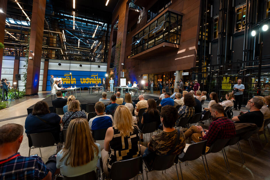 Zdjęcie przedstawia wnętrze nowoczesnego budynku, w którym odbywa się konferencja lub publiczna dyskusja. W centrum znajduje się podwyższona scena, na której siedzą prelegenci przy stołach. Na scenie widoczne są także tablice prezentacyjne i sprzęt do nagrywania. W tle sceny znajduje się duży ekran z napisem UKRAINA  w żółto-niebieskich barwach, co może sugerować, że temat dyskusji jest związany z Ukrainą. Przed sceną siedzi licznie zgromadzona publiczność na plastikowych krzesłach, zwrócona twarzą w stronę sceny. W pierwszym rzędzie można zauważyć osoby różnych płci i wieku, skupione na wydarzeniu. W głębi po lewej stronie widoczna jest kamera z operatorem, co wskazuje na rejestrowanie lub transmisję na żywo wydarzenia.