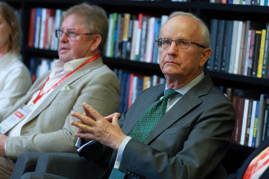 Na zdjęciu widoczni są dwaj starsi mężczyźni siedzący przed półką z książkami. Mężczyzna na pierwszym planie ma na sobie szary garnitur, zielony krawat w wzór pawich oczu oraz okulary, a jego dłonie są złożone w geście przypominającym zaangażowaną rozmowę lub debatę.