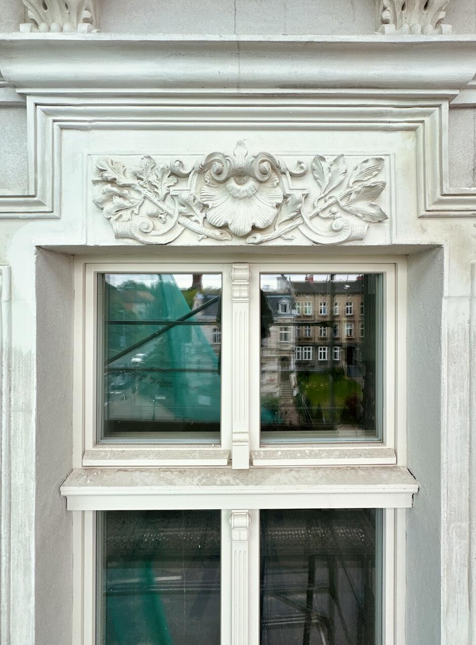 Zdjęcie przedstawia okno z ozdobnym nadprożem na fasadzie budynku. Nadproże jest zdobione dekoracyjnymi płaskorzeźbami roślinnymi, w tym dużym kwiatem centralnym oraz liśćmi po bokach. Okno składa się z dwóch dużych szyb przedzielonych pionowym elementem z delikatnymi zdobieniami. Ramy okna są białe, co harmonizuje z ozdobnym nadprożem i resztą fasady. W oknie odbija się otoczenie, w tym inne budynki i zielone siatki ochronne na rusztowaniach, wskazujące na trwające prace renowacyjne. Całość utrzymana jest w klasycznym stylu z dużą dbałością o detale architektoniczne.