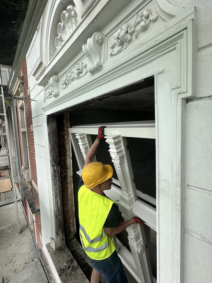 Zdjęcie przedstawia pracownika budowlanego podczas renowacji fasady budynku. Mężczyzna, ubrany w żółty kask ochronny i jaskrawą kamizelkę odblaskową, montuje ozdobne okno. Widać, że okno ma bogato zdobione kolumny i detale architektoniczne, podobne do tych widocznych na poprzednich zdjęciach. Pracownik używa czerwonych rękawic roboczych, aby ostrożnie ustawić elementy okna w odpowiednim miejscu.