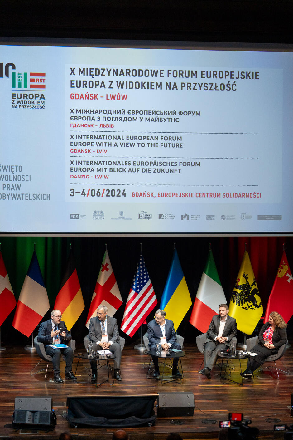 Na zdjęciu widzimy panel dyskusyjny podczas X Międzynarodowego Forum Europejskiego Europa z widokiem na przyszłość  w Gdańsku. W tle znajduje się ekran z informacjami o wydarzeniu oraz flagi różnych państw, a na scenie siedzi pięciu mężczyzn i jedna kobieta, uczestniczący w debacie