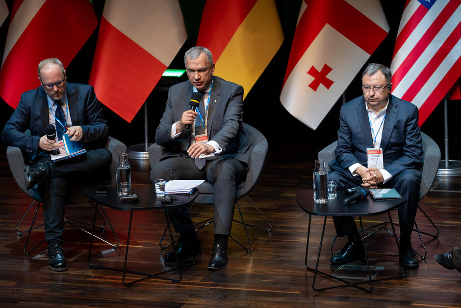 Na zdjęciu widzimy trzech mężczyzn siedzących na scenie podczas panelu dyskusyjnego, każdy z mikrofonem w ręku. W tle znajdują się flagi różnych państw, a na stolikach przed uczestnikami stoją butelki z wodą i szklanki.