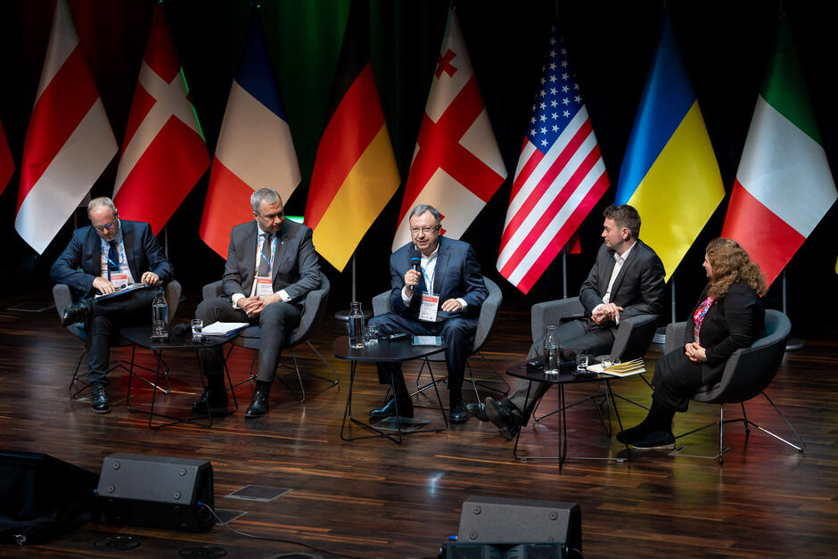 Na zdjęciu znajduje się panel dyskusyjny z udziałem pięciu osób: czterech mężczyzn i jednej kobiety, siedzących na scenie i rozmawiających. W tle widoczne są flagi różnych państw, w tym Polski, Danii, Francji, Niemiec, Gruzji, Stanów Zjednoczonych, Ukrainy, Włoch, Flandrii i kolejna flaga Polski.