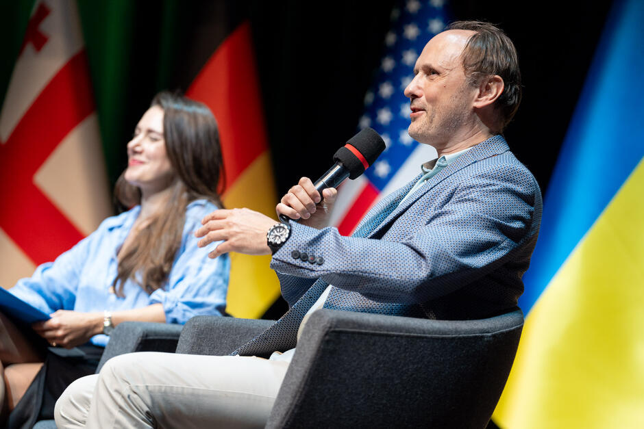 Na zdjęciu widzimy mężczyznę w jasnej marynarce, siedzącego na krześle i trzymającego mikrofon, obok kobiety w niebieskiej koszuli, również siedzącej na krześle. W tle widoczne są flagi różnych krajów, w tym Niemiec, Stanów Zjednoczonych, Gruzji i Ukrainy.
