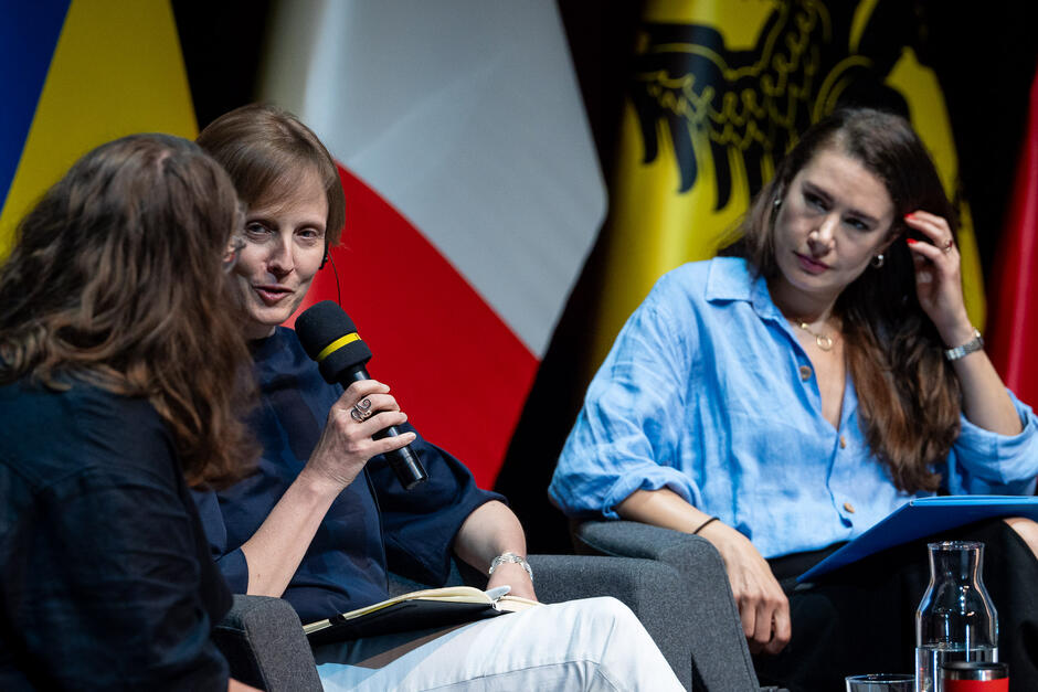 Na zdjęciu widzimy trzy kobiety uczestniczące w panelu dyskusyjnym, z których jedna trzyma mikrofon i przemawia, podczas gdy dwie pozostałe słuchają. W tle widoczne są flagi różnych krajów, w tym Polski, Ukrainy i Włoch.