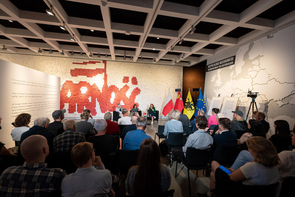 Trzy osoby - dwie kobiety i mężczyzna siedzą na fotelach i rozmawiają. Przed nimi na krzesłach siedzi publiczność. Na ścianie w sali, w której odbywa się spotkanie autorskie widać napis Solidarność . Obok stoją flagi Polski, Gdańska, Kaszub i Unii Europejskiej.