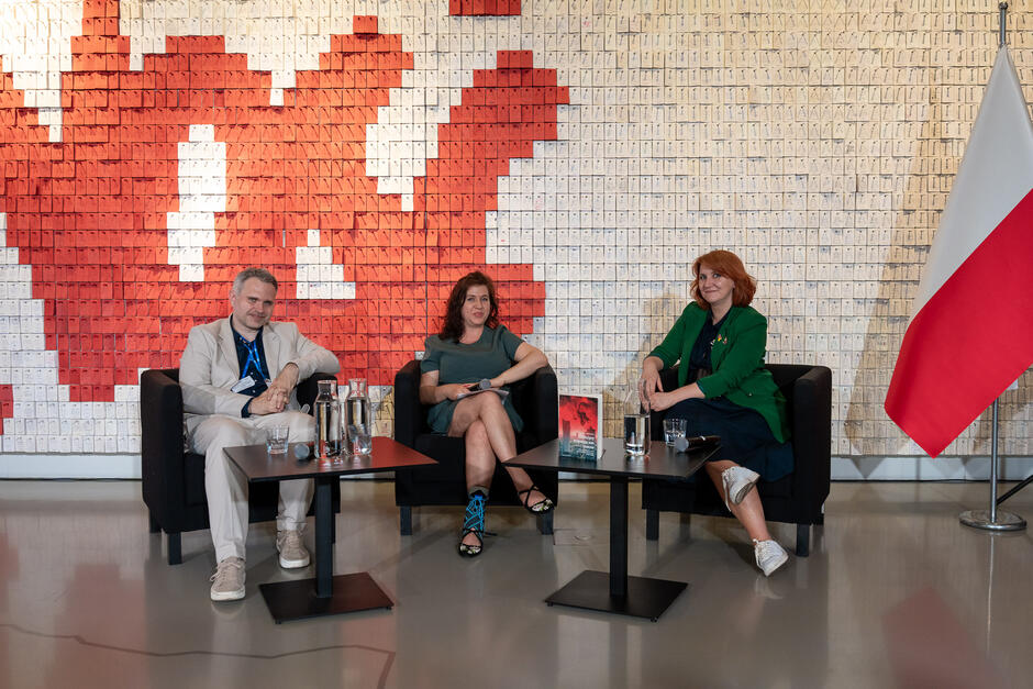 Trzy osoby, dwie kobiety i mężczyzna, siedzą na fotelach i rozmawiają podczas spotkania autorskiego. Za nimi ściana z napisem Solidarność .