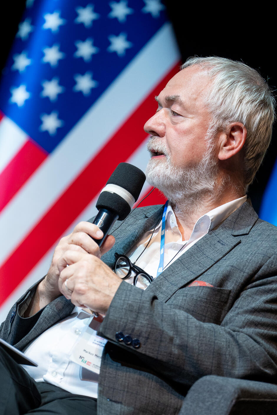 Na zdjęciu widzimy starszego mężczyznę z siwymi włosami i brodą, który trzyma mikrofon i przemawia, siedząc na tle flagi Stanów Zjednoczonych. Mężczyzna jest elegancko ubrany w szary garnitur i białą koszulę, a na szyi ma identyfikator konferencyjny.