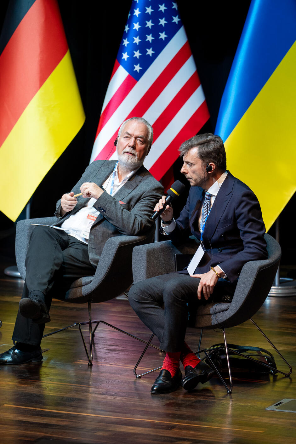 Na zdjęciu widzimy dwóch mężczyzn siedzących na scenie przed flagami Niemiec, Stanów Zjednoczonych i Ukrainy. Starszy mężczyzna po lewej stronie ma siwe włosy i brodę, trzyma w ręku mikrofon, a młodszy mężczyzna po prawej stronie, w ciemnym garniturze i czerwonych skarpetkach, również trzyma mikrofon i przemawia.