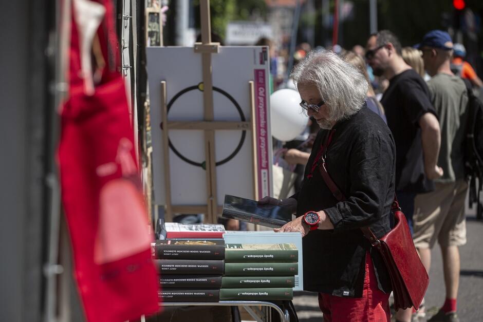 Na zdjęciu widzimy mężczyznę z długimi, siwymi włosami i w okularach, przeglądającego książki na stoisku podczas plenerowej imprezy. W tle znajduje się grupa ludzi spacerujących wzdłuż ulicy