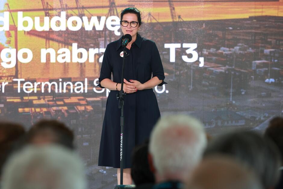 Na zdjęciu widzimy kobietę stojącą przy mikrofonie na tle napisu informującego o budowie nowego nabrzeża T3. Uroczystość związana jest z rozbudową terminala kontenerowego Baltic Hub.