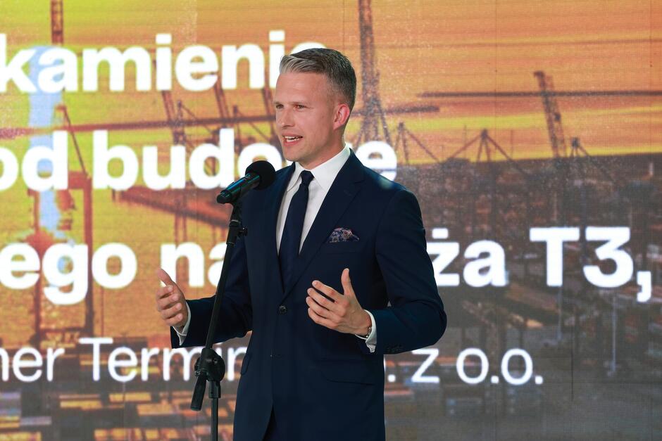 Na zdjęciu widzimy mężczyznę w garniturze przemawiającego przy mikrofonie na tle napisu dotyczącego budowy nabrzeża T3. Uroczystość odbywa się w związku z rozbudową terminala kontenerowego Baltic Hub.