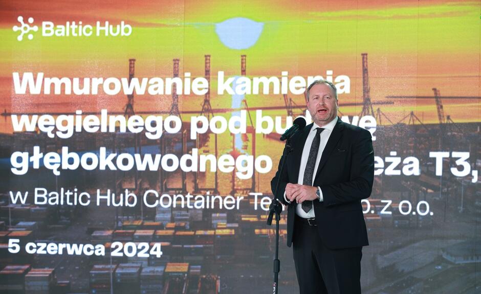 Na zdjęciu widzimy mężczyznę w garniturze przemawiającego przy mikrofonie na tle napisu informującego o wmurowaniu kamienia węgielnego pod budowę głębokowodnego nabrzeża T3. Uroczystość odbyła się 5 czerwca 2024 roku w Baltic Hub Container Terminal.