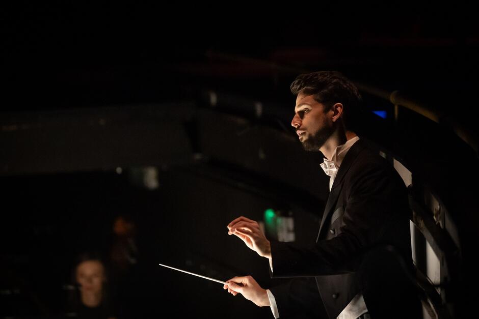 Zdjęcie przedstawia dyrygenta w eleganckim garniturze, który z koncentracją prowadzi orkiestrę. Mężczyzna trzyma batutę i jest oświetlony subtelnym światłem