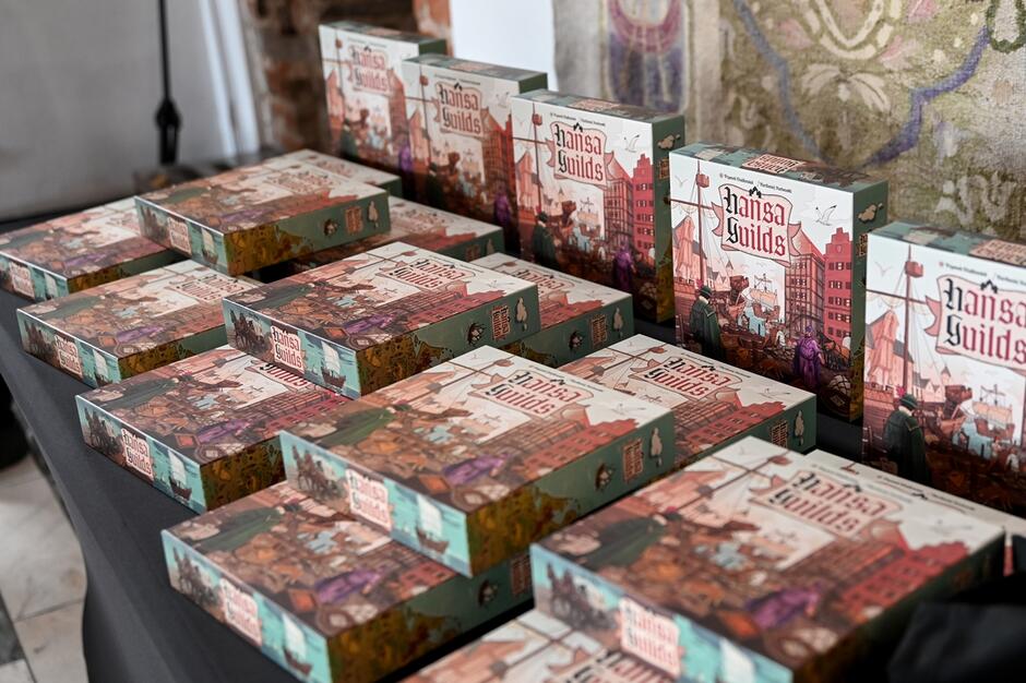 Na zdjęciu widzimy wiele pudełek gry planszowej Hansa Guilds  ułożonych na stole. Każde pudełko ma kolorową ilustrację przedstawiającą średniowieczne miasto z portem, co nawiązuje do tematyki gry związanej z hanzeatyckimi kupcami i gildiami.