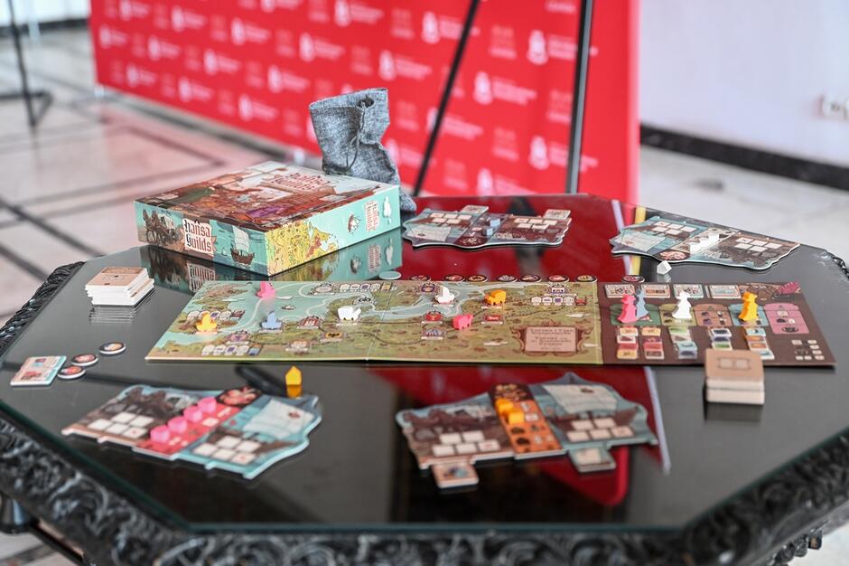 Na zdjęciu widzimy rozłożoną grę planszową Hansa Guilds  na eleganckim stole. Elementy gry, w tym plansza, karty, żetony i figurki, są starannie ułożone, a w tle znajduje się czerwone tło z logo, sugerujące, że jest to prezentacja lub konferencja prasowa związana z grą.