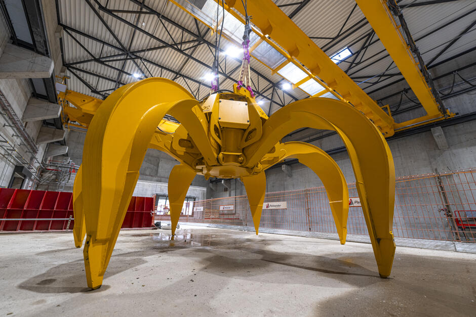 Na zdjęciu widać dużą, żółtą ładowarkę przemysłową, która znajduje się wewnątrz hali. Maszyna ma kilka masywnych ramion i jest zawieszona na żurawiu, przygotowana do podjęcia ciężkich zadań w obszarze przemysłowym