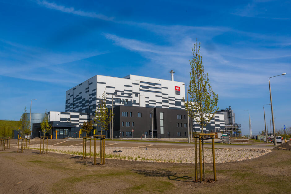 Na zdjęciu znajduje się nowoczesny budynek przemysłowy oznaczony jako Port Czystej Energii . Budynek ma biało-czarne, geometryczne elewacje i jest otoczony nowo nasadzonymi drzewami oraz uporządkowanym terenem