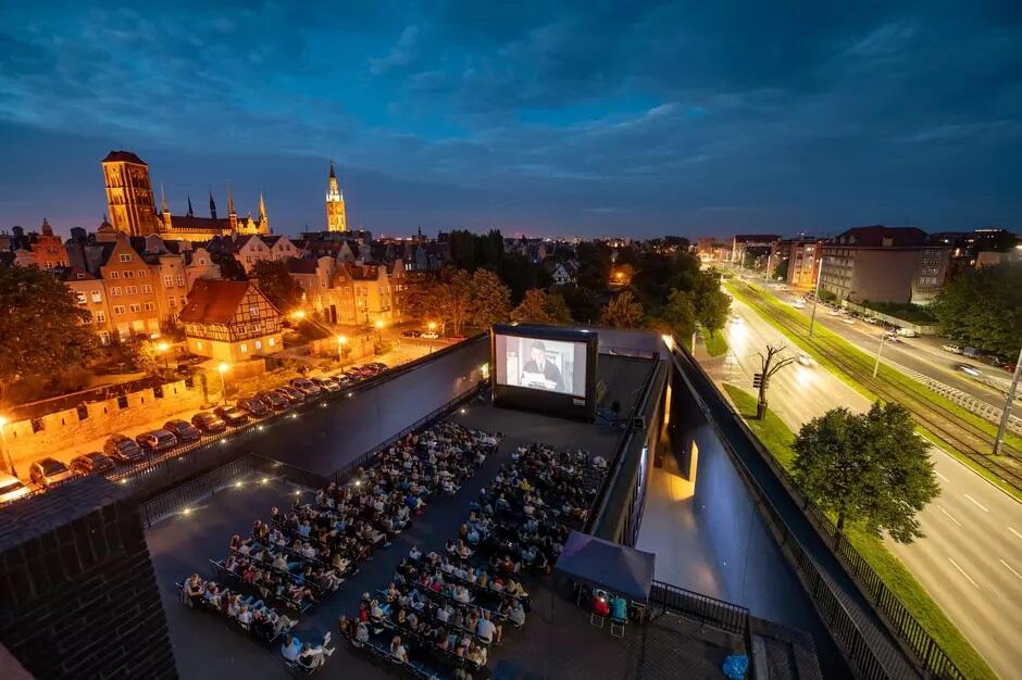 Na zdjęciu widać kino plenerowe na dachu budynku, gdzie ludzie oglądają film pod gołym niebem. W tle rozciąga się widok na urokliwe, oświetlone nocą miasto z historycznymi budynkami i kościołami, co tworzy malowniczą scenerię
