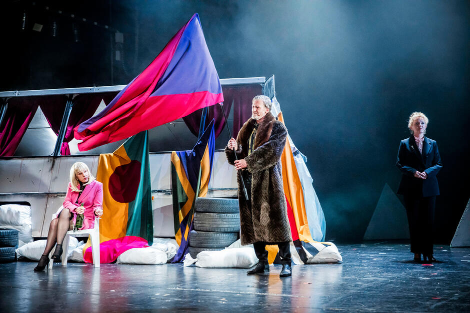 Zdjęcie przedstawia scenę teatralną, na której znajduje się trzech aktorów w różnych strojach. Mężczyzna w futrze trzyma flagę, kobieta w różowym kostiumie siedzi na krześle z butelką w dłoni, a trzecia osoba stoi w oddali w ciemnym garniturze