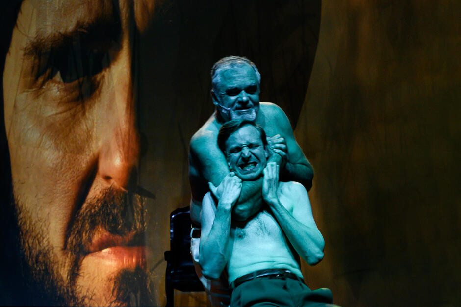 Zdjęcie przedstawia dwóch półnagich mężczyzn w dramatycznej scenie, gdzie jeden z nich dusi drugiego, wyrażając silne emocje. W tle widać duży zbliżenie twarzy trzeciej osoby