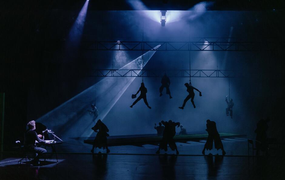 Zdjęcie przedstawia scenę teatralną w ciemnym, tajemniczym oświetleniu, gdzie postacie unoszą się w powietrzu na linach, tworząc dramatyczny efekt. Po lewej stronie widać osobę siedzącą przy biurku