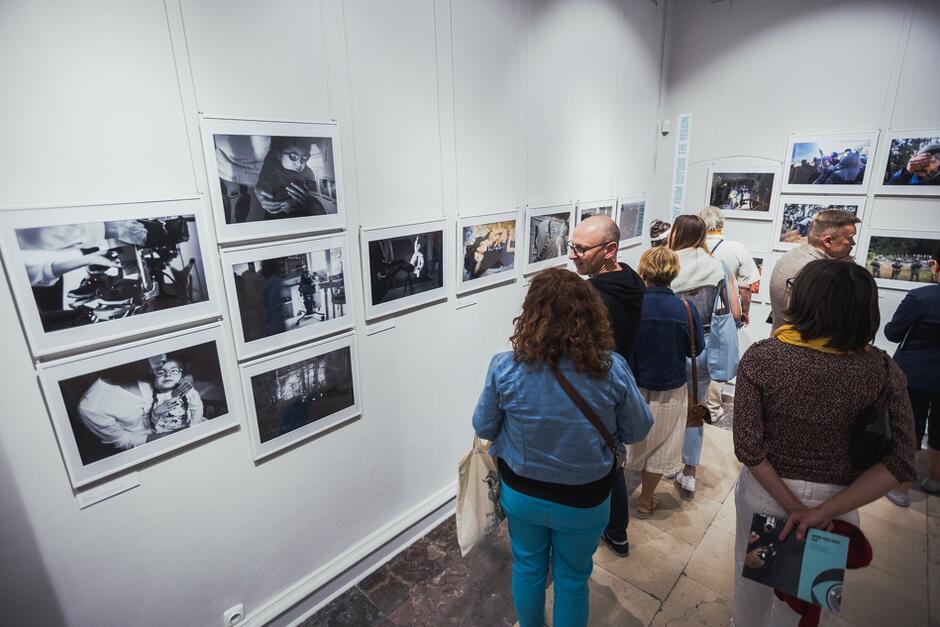 Na zdjęciu widać grupę ludzi oglądających czarno-białe fotografie na wystawie w galerii sztuki. Ludzie są skupieni na zdjęciach, rozmawiają między sobą i poruszają się po przestrzeni wystawienniczej