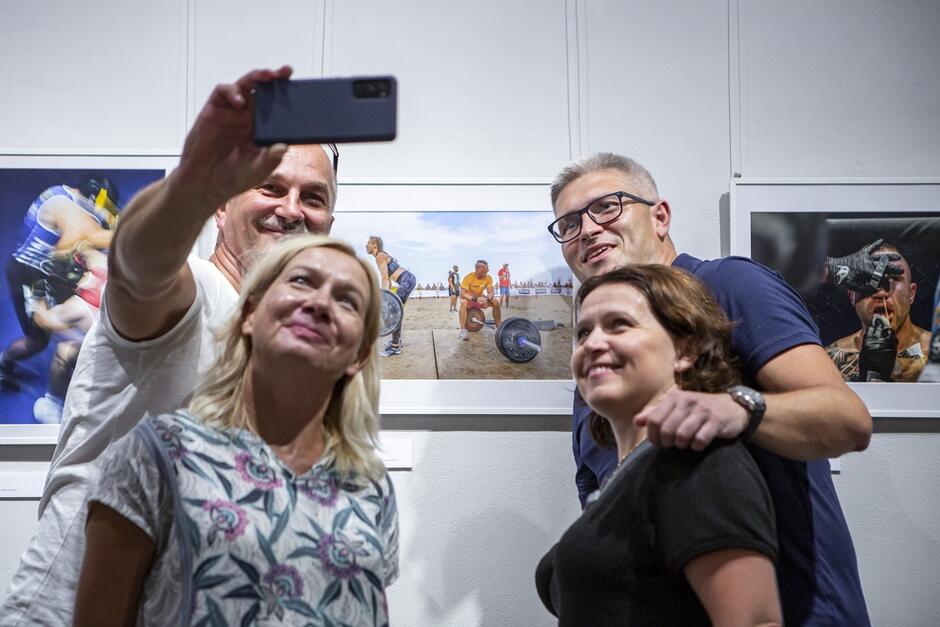 Na zdjęciu widać cztery osoby robiące sobie selfie przed kolorowymi fotografiami sportowymi zawieszonymi na ścianie w galerii sztuki. Wszyscy wyglądają na zadowolonych i uśmiechają się do kamery, tworząc radosną atmosferę