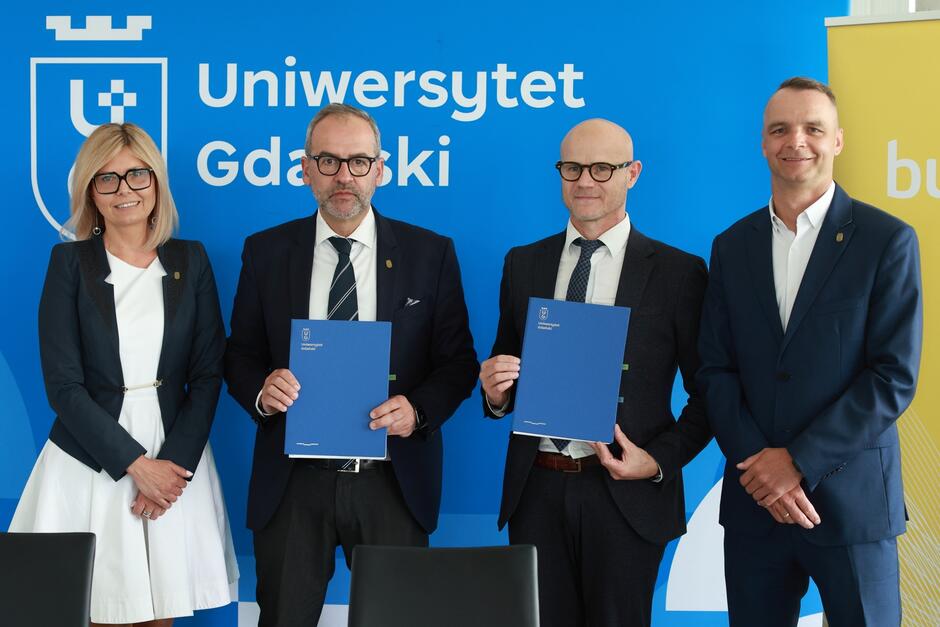 Zdjęcie przedstawia cztery osoby stojące na tle niebieskiej ścianki z logo i nazwą Uniwersytetu Gdańskiego. Dwóch mężczyzn w środku trzyma niebieskie teczki z logo uczelni, sugerujące podpisanie lub prezentację dokumentów, podczas gdy pozostałe osoby stoją po bokach i uśmiechają się do kamery.