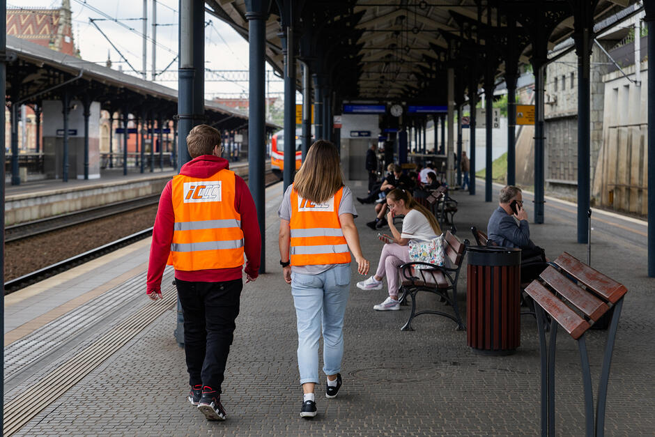 Dwie osoby w pomarańczowych kamizelkach spacerują po peronie kolejowym. Na ławkach siedzą pasażerowie.