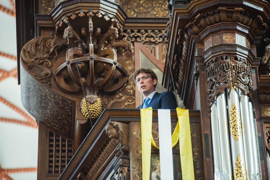 Zdjęcie przedstawia mężczyznę stojącego na ozdobnej, drewnianej ambonie wewnątrz bogato zdobionego kościoła. Mężczyzna ma na sobie garnitur i krawat, a ambona ozdobiona jest złoconymi elementami oraz żółtymi i białymi wstęgami.