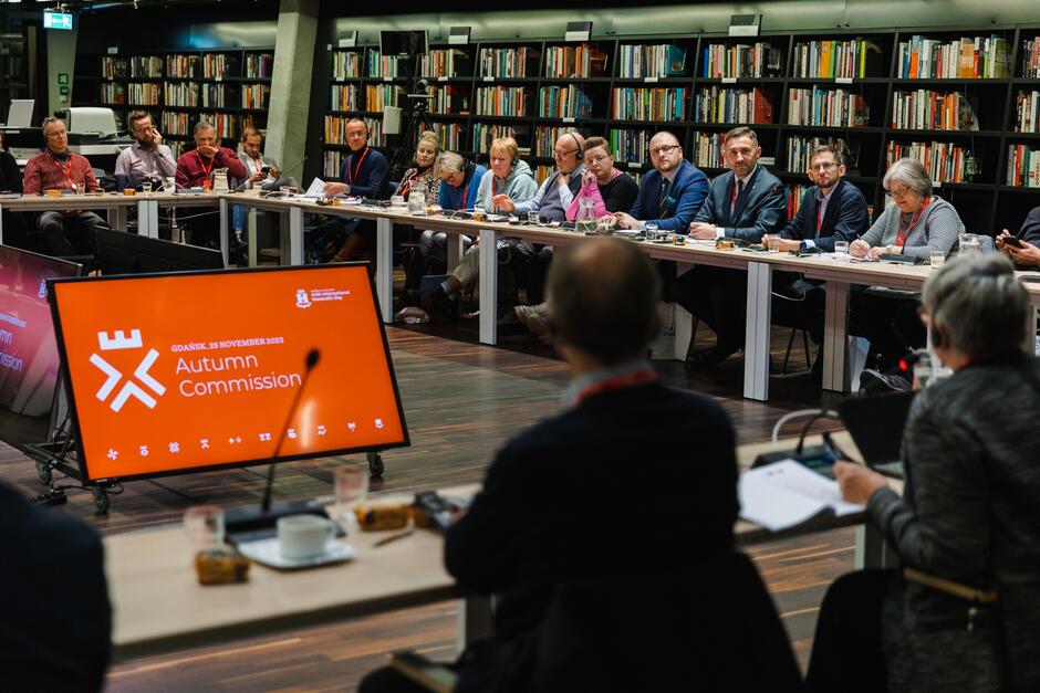 Zdjęcie przedstawia spotkanie w sali konferencyjnej, w której uczestniczy kilkanaście osób siedzących przy prostokątnych stołach ustawionych w kształt litery U. Na pierwszym planie widoczny jest ekran z napisem Autumn Commission  i datą  25 November 2023,  sugerując, że jest to oficjalne wydarzenie odbywające się w Gdańsku.