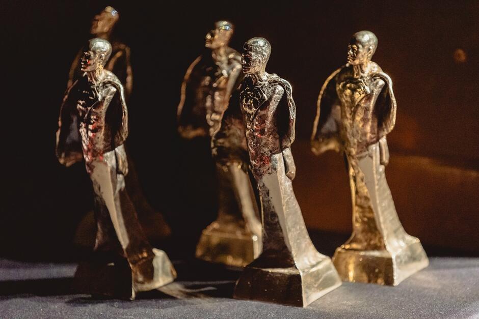 Na zdjęciu widzimy grupę statuetek przedstawiających postacie ludzkie, wykonane z metalu i umieszczone na ciemnym tle. Statuetki te najprawdopodobniej służą jako nagrody, które mają być wręczone podczas uroczystej ceremonii.