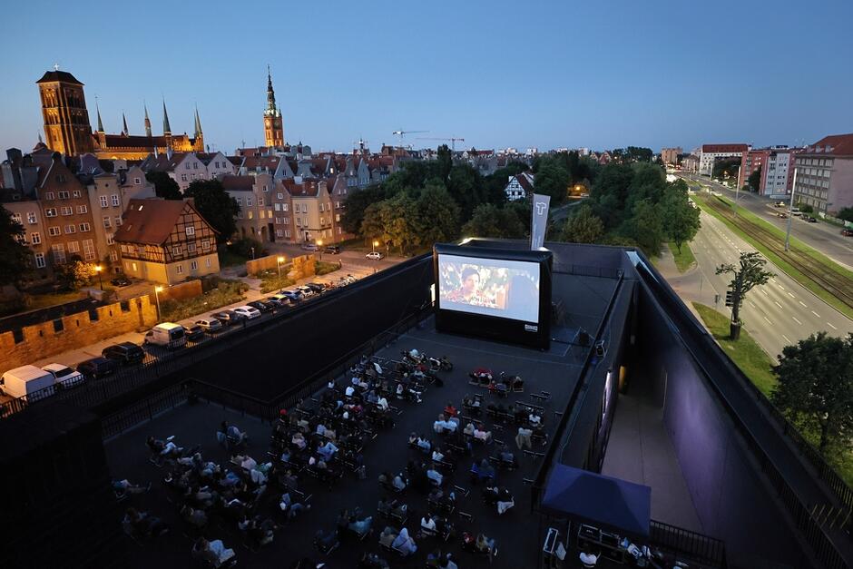 Na zdjęciu widzimy wieczorny seans filmowy na świeżym powietrzu, odbywający się na dachu budynku w Gdańsku. W tle widać malowniczy widok na zabytkową część miasta, z wyraźnie widocznymi wieżami kościołów i ratusza, co tworzy unikalną atmosferę dla widzów