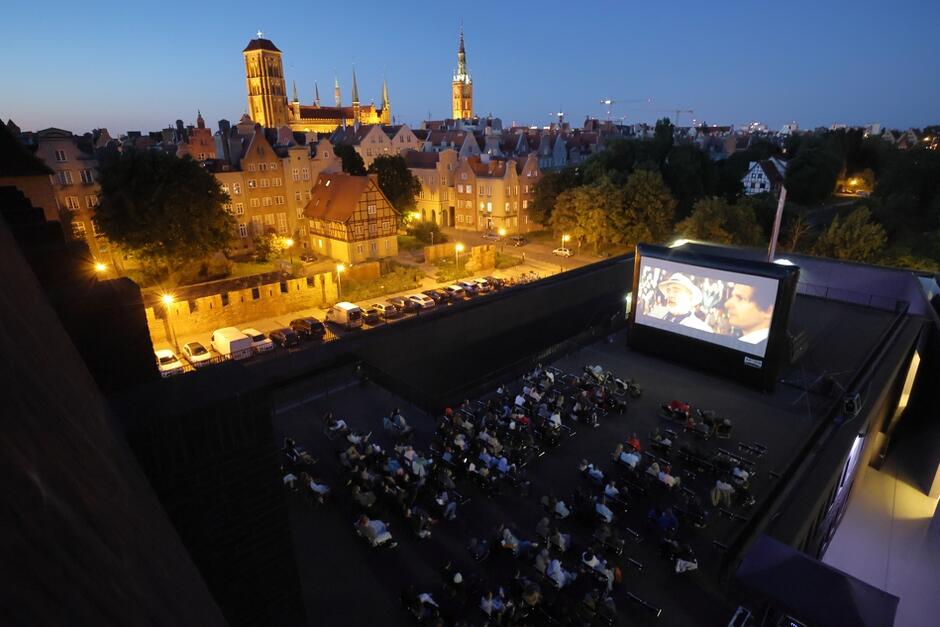 Na zdjęciu widzimy wieczorny seans filmowy na świeżym powietrzu, odbywający się na dachu budynku w Gdańsku. W tle widać malowniczy widok na zabytkową część miasta, z wyraźnie widocznymi wieżami kościołów i ratusza, co tworzy unikalną atmosferę dla widzów