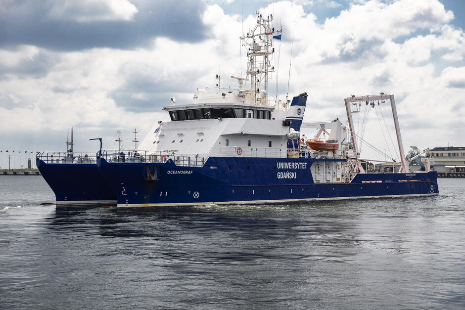 Na zdjęciu widać nowoczesny katamaran badawczy OCEANOGRAF  należący do Uniwersytetu Gdańskiego, płynący po spokojnych wodach portu. Statek jest pomalowany na kolor niebieski z białą nadbudówką, a w tle widoczne jest nabrzeże z masztami flagowymi oraz budynkami portowymi.